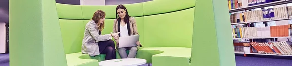 Zwei Frauen besprechen etwas an einem Laptop und sitzen auf einem grünen Sofa