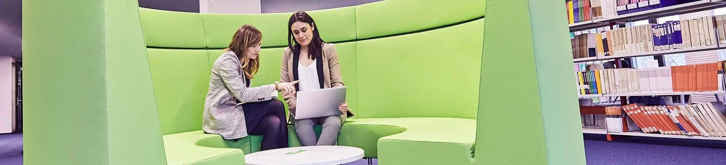 Zwei Frauen besprechen etwas an einem Laptop und sitzen auf einem grünen Sofa